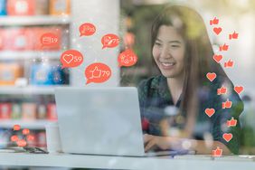 社交媒体概念，亚洲女性用笔记本电脑检查社交网络应用程序的数量，喜欢，评论，人和最喜欢的图标在咖啡店玻璃旁边的桌子上