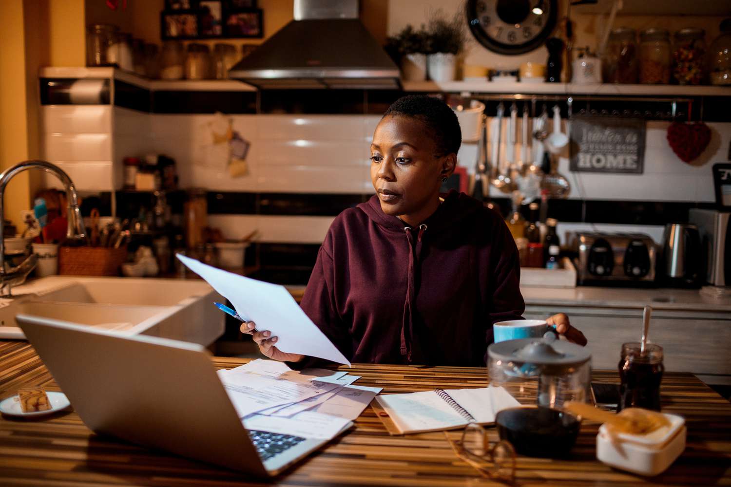 一名妇女坐在厨房桌子旁，面前是一台打开的笔记本电脑，她正专注地看着手里的一份文件