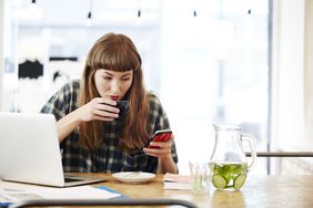 Young woman checking her phone while drinking coffee in front of her laptop