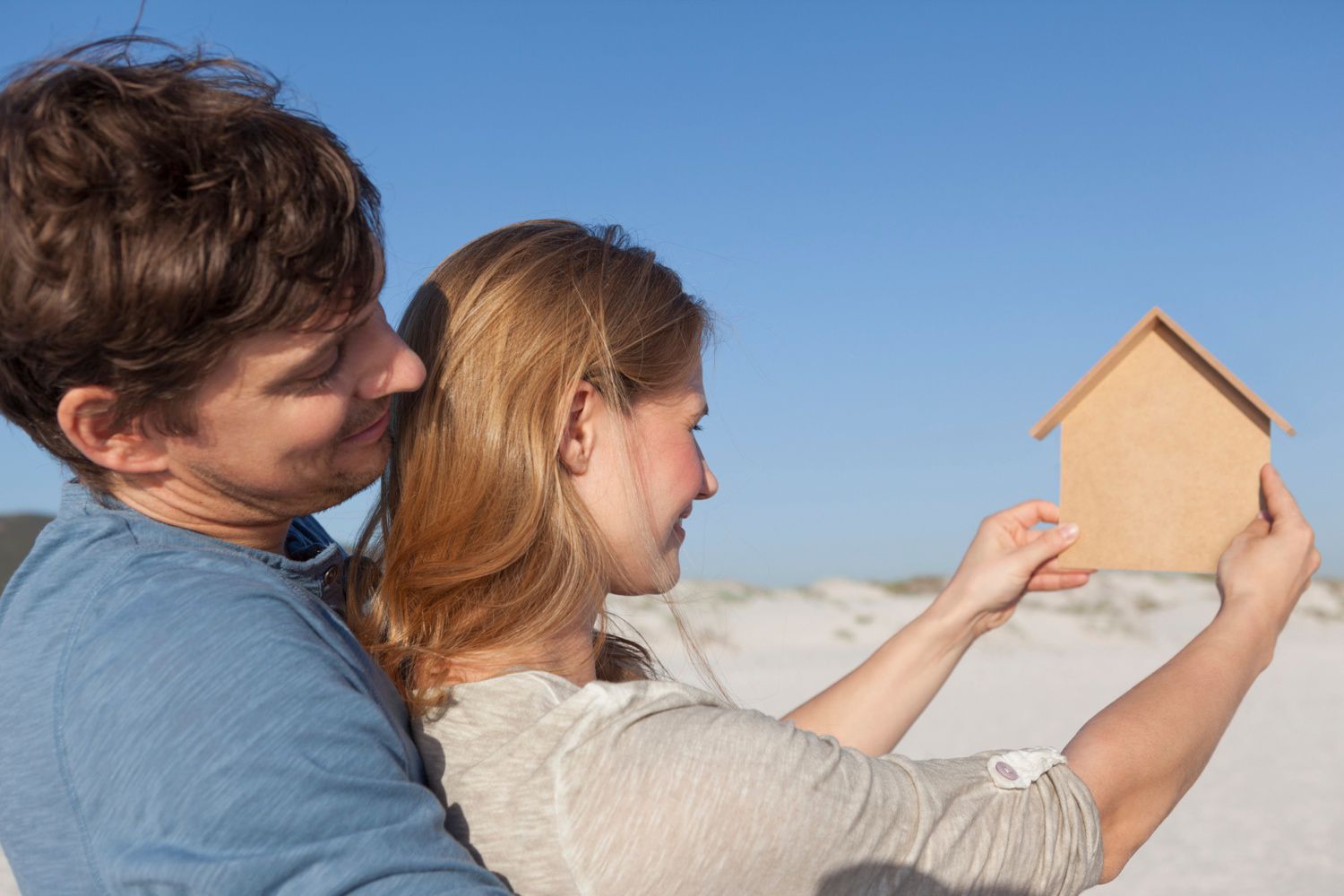 一对夫妇计划在未来的海滨别墅站在海滩上看着一个纸板剪出的房子。