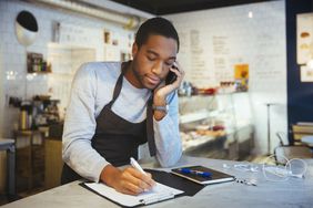 一个年轻的黑人商人靠在柜台上讲电话，在笔记本上记笔记。