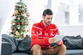 穿着红白相间的节日毛衣的人拿着平板电脑坐在圣诞树前