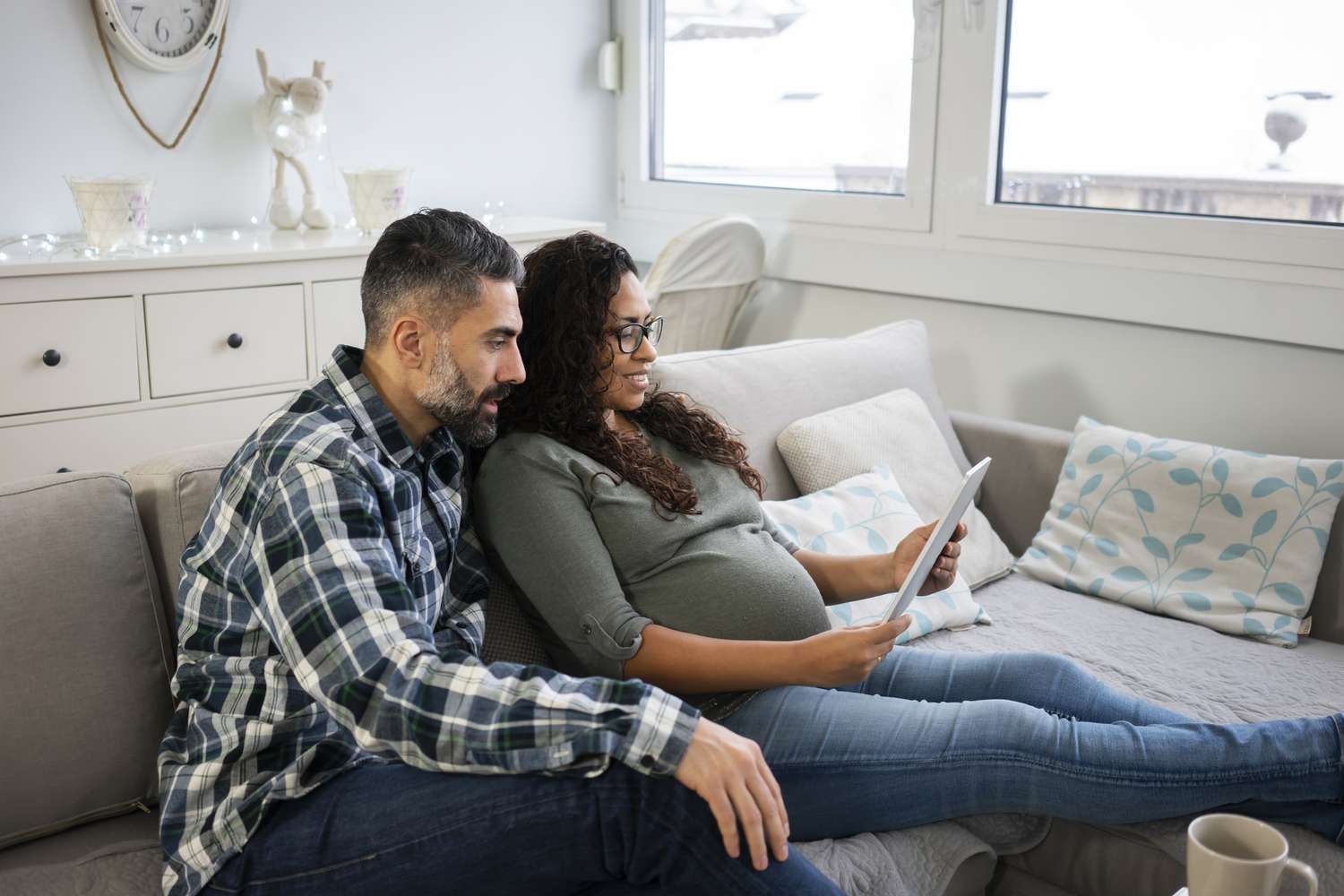 一个留着胡子的男人和一个孕妇坐在沙发上,看着他们的预算在平板电脑上”class=