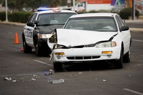 一辆警车停在一辆发生事故的白色轿车后面，乘客侧严重受损，周围有破碎的玻璃和塑料。