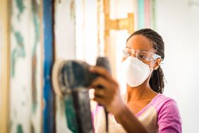 一名女子戴着口罩和护目镜，正在用电动砂光机打磨墙壁