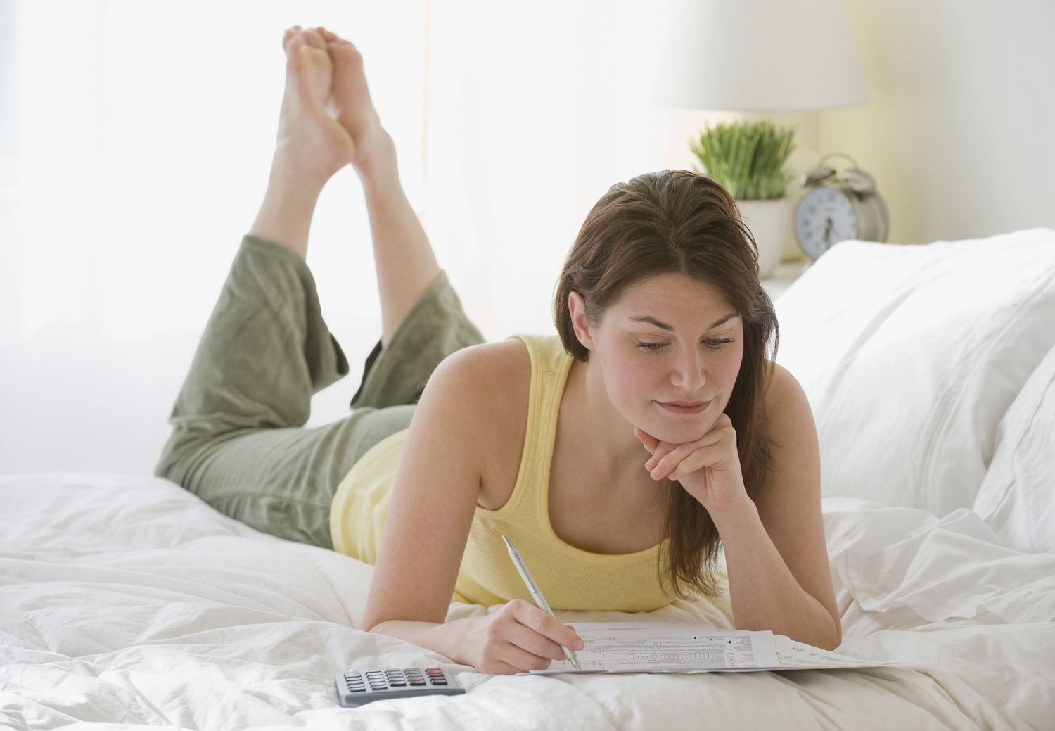 女人躺在床上使用一个计算器,因为她完成联邦税收回报”class=