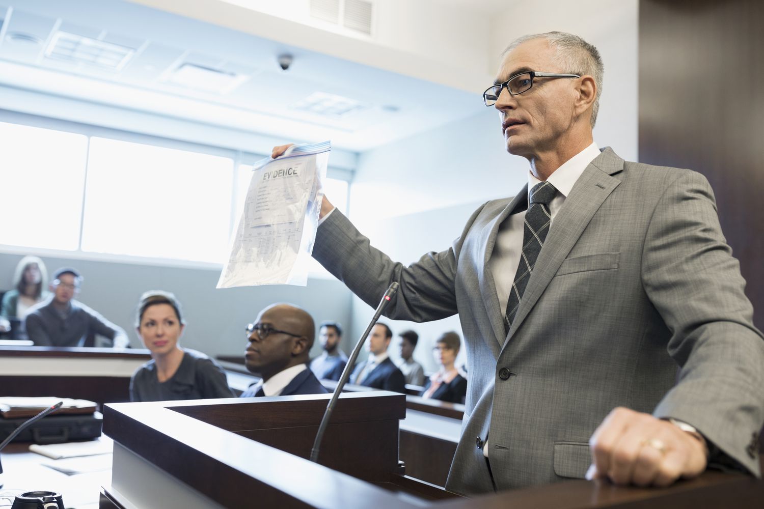 一位头发花白的绅士律师站在法庭的讲台上，右手拿着一份标有“证据”的文件。
