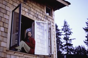 一个穿着红色法兰绒的女人坐在一间小屋敞开的窗户上，带着极度满足的表情向外凝视。