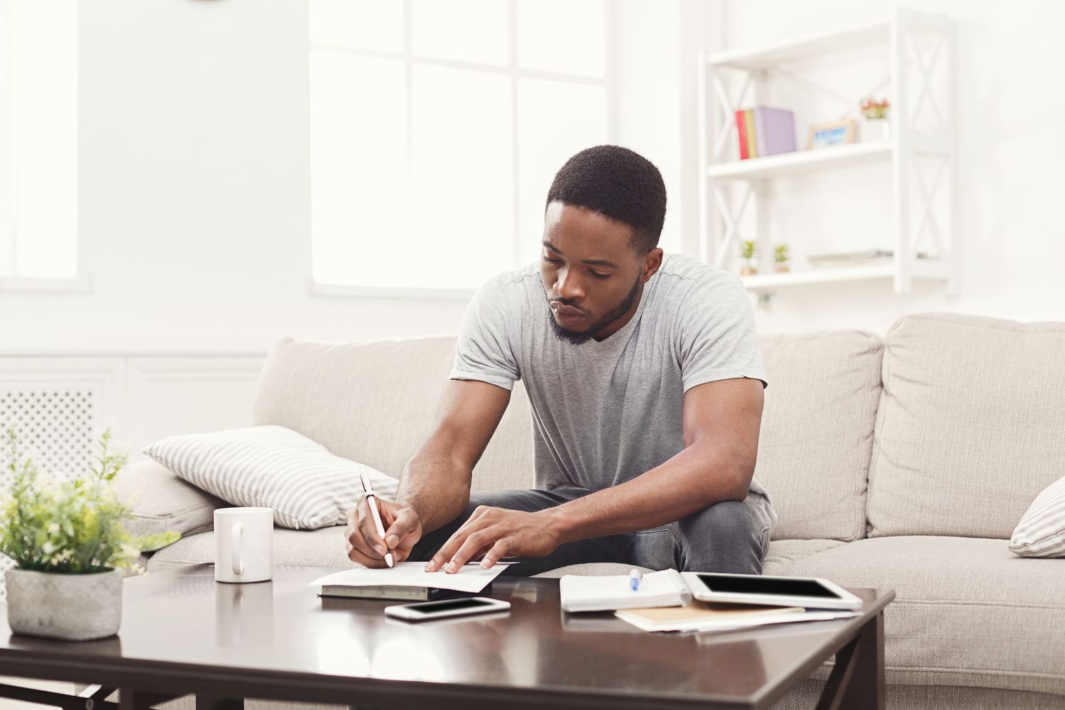 一个年轻人坐在沙发上，拿着笔记本电脑和手机在咖啡桌旁写作