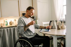 纹身的成熟男子阅读邮件，同时在轮椅上拿着咖啡杯在家里
