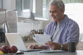白发男子在笔记本电脑上工作，一手拿着纸质财务报表