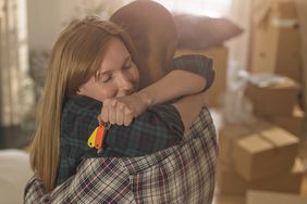 A young couple hugs in a room full of boxes in their new home.