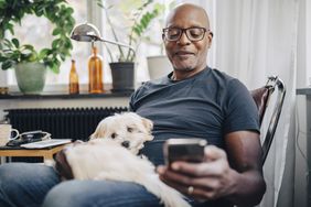 微笑的退休高级男性在家里的房间里和狗坐在一起用智能手机。