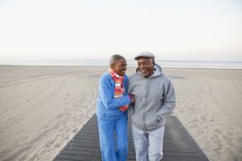老夫妇手挽着手在沙滩上散步，一起欢笑。＂width=