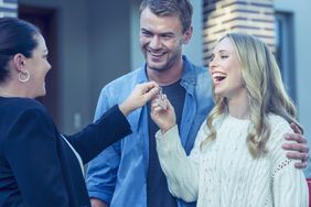 Couple standing in front of a new home with agent. They are standing next to a for sale sign with a sold sticker.