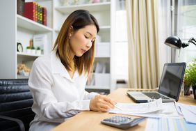 一名女子坐在办公室的桌子前，拿着铅笔检查一份文件，面前是一台打开的笔记本电脑，前景上有一个计算器