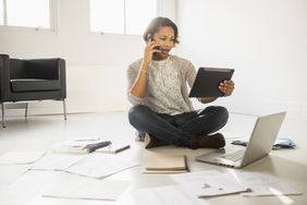 一名女子一边打电话，一边看着平板电脑，周围的地板上放着纸、笔记本电脑和笔记本电脑