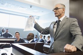一位头发花白的绅士律师站在法庭的讲台上，右手拿着一份标有“证据”的文件。