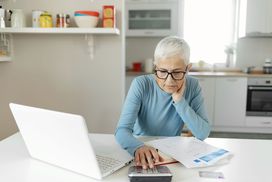 女人在蓝色上衣灰色短发使用计算器和笔记本在她的厨房。”width=
