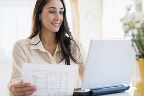 一位快乐的女士拿着打印好的预算，看着她的支票账户余额，对着笔记本电脑微笑