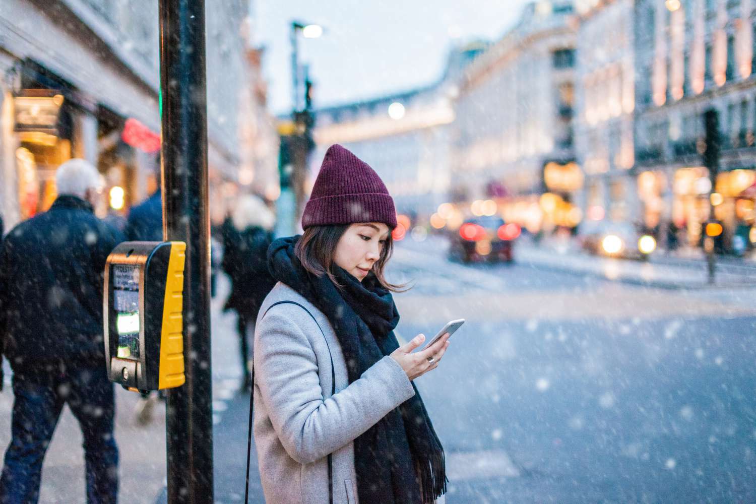 在下雪的天气里，一名戴着帽子的女子站在城市的角落里看手机＂class=