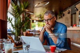 一名男子拿着笔记本电脑坐在咖啡桌旁，看起来若有所思＂width=