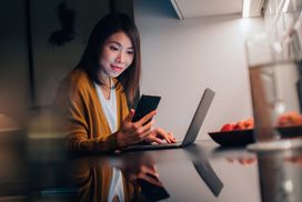 年轻的亚洲妇女看智能手机与笔记本电脑在厨房工作时在晚上在家”width=