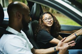 Teenage girl in drivers seat talks to driving instructor in car.