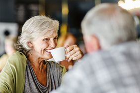 一头灰色短发的女人微笑着从咖啡杯里喝了一口。一个男人坐在她对面，背对着镜头。他在前景，焦距不够。