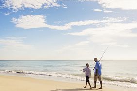 一对父子肩上扛着鱼竿，沿着海滩散步。