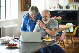 老人在早餐吧台吃炒鸡蛋，女人把手放在他的肩膀上看笔记本电脑，一对夫妇在早餐时讨论家庭财务＂width=