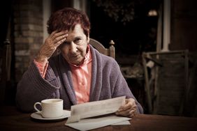 一位穿着长袍的老妇人一边喝着咖啡，一边端详着医药费账单。