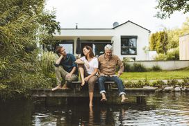 两名男子和一名女子坐在房子外靠近水面的甲板上＂width=