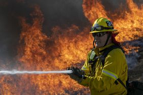 A firefighter with a hose, fire raging behind him