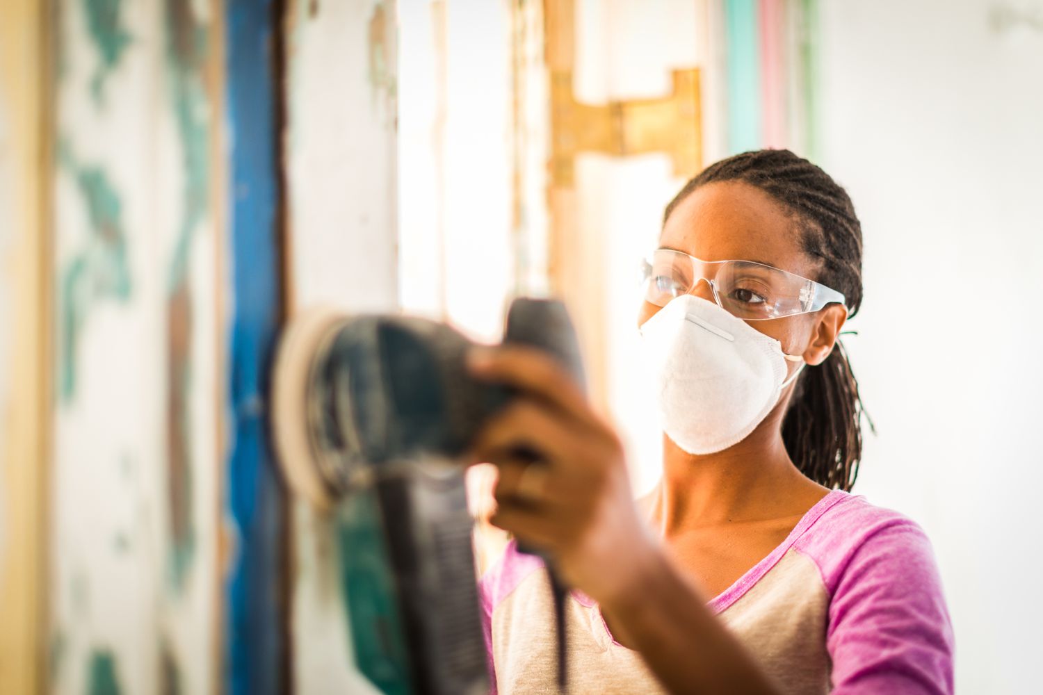 一名女子戴着口罩和护目镜，正在用电动砂光机打磨墙壁＂class=