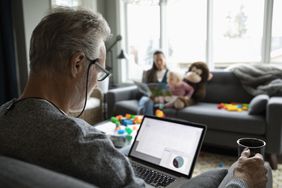 老人在客厅用笔记本电脑喝咖啡，背景是家人