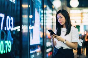 在市中心金融区，一名女子通过股市显示屏查看智能手机上的股票价格