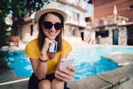 一名女子坐在波光粼粼的游泳池附近的阴凉处，手里拿着信用卡，用着智能手机＂width=