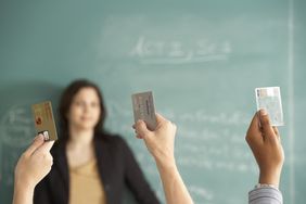 三名学生举起手，每人拿着一张信用卡，而在背景中，一位老师站在黑板前，一脸困惑地看着。