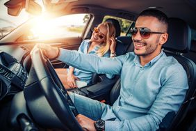 一个男人开着一辆车，副驾驶座上坐着一个女人。两人都戴着墨镜，脸上挂着灿烂的笑容，阳光从副驾驶侧的窗户照进来。