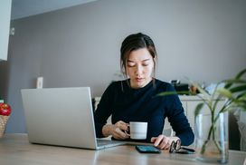 年轻女子一边喝咖啡一边用笔记本电脑和手机工作＂width=