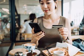 一名女子在餐厅用餐时，用智能手机管理网上银行，手持信用卡进行移动支付