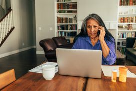 一个女人坐在厨房桌子旁，一边用笔记本电脑付账单，一边打电话＂width=