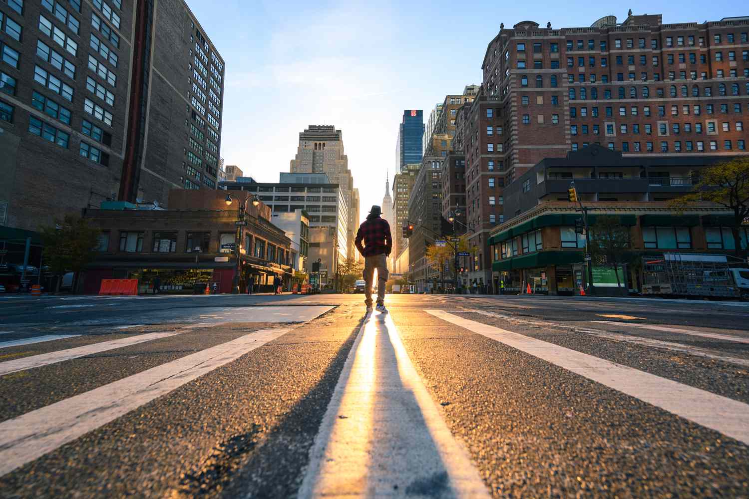 一个穿着格子夹克，戴着软呢帽的人站在纽约一个十字路口的中央，身后街道上的阳光照亮了他