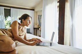 一位年轻女子坐在床上，面前是一台打开的笔记本电脑，她双手放在键盘上，专注地看着屏幕