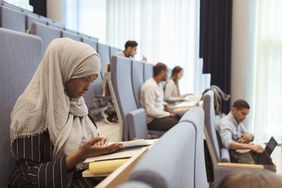 在一所大学的讲堂里，一名戴着头巾的严肃女子正在看书，背景中还有其他学生。