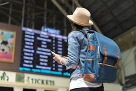 在一个机场候机楼里，一位年轻的背包女游客抬头看着头顶上的航班信息板。＂width=
