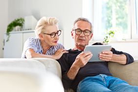 老人坐在家里客厅的沙发上，给妻子看平板电脑上的东西。