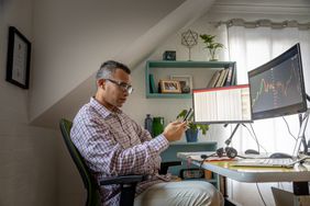 一名男子坐在桌子前，桌子上放着一台笔记本电脑和两个显示图表的电脑显示器。