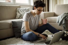 穿着灰色t恤的人坐在地板上，一边喝咖啡一边在电脑上工作＂width=
